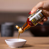 Hand pouring a liquid from a bottle labeled 'SIAM' fish sauce into a small white bowl on a wooden surface.
